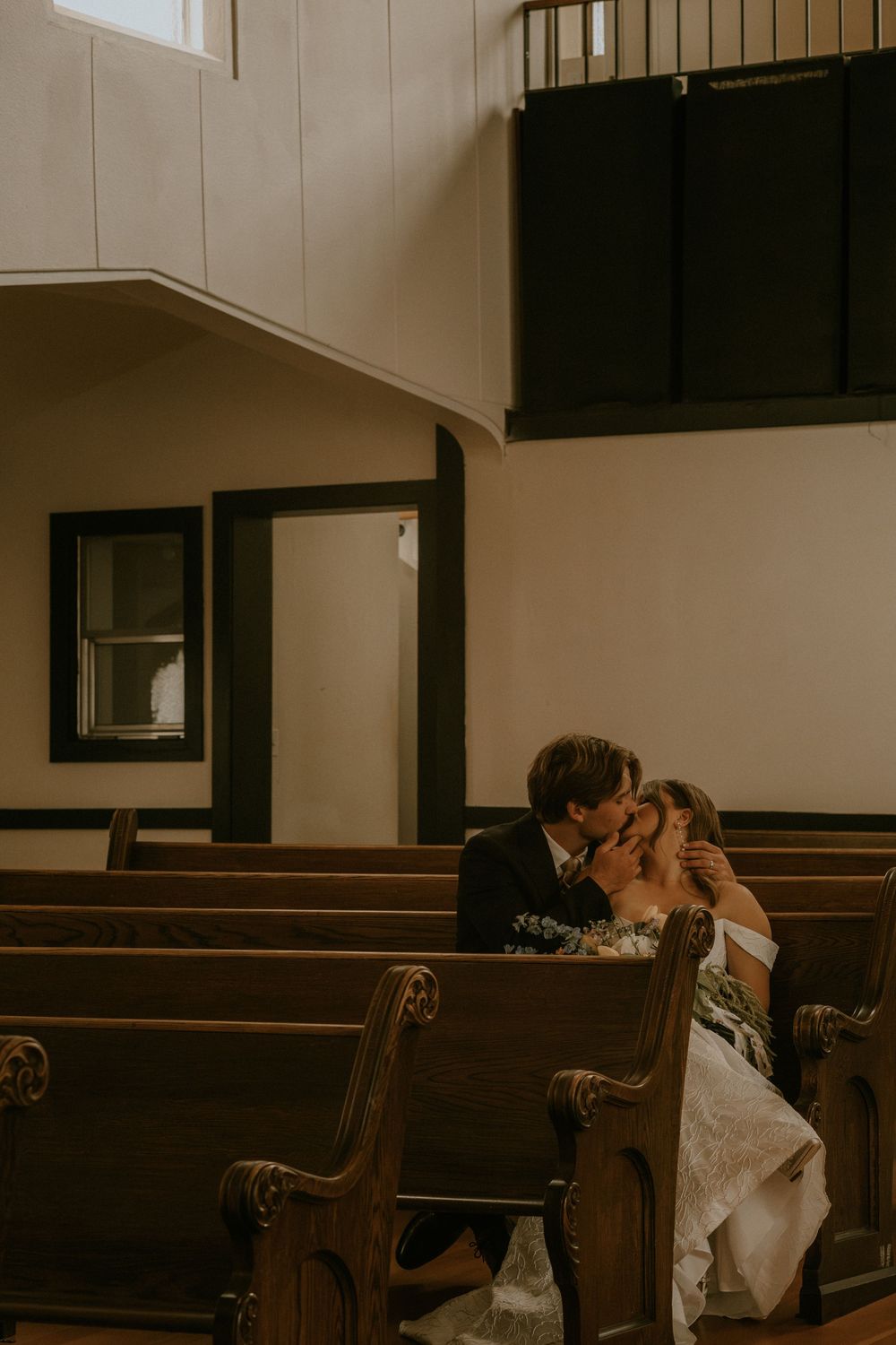 Bride in a white lace gown and groom in a dark suit share a kiss on a wooden church pew in a quiet, sunlit chapel.