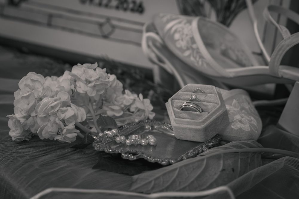 Black and white wedding details showing an open ring box with rings, pearl jewelry, white flowers, and embellished bridal shoes on a table.