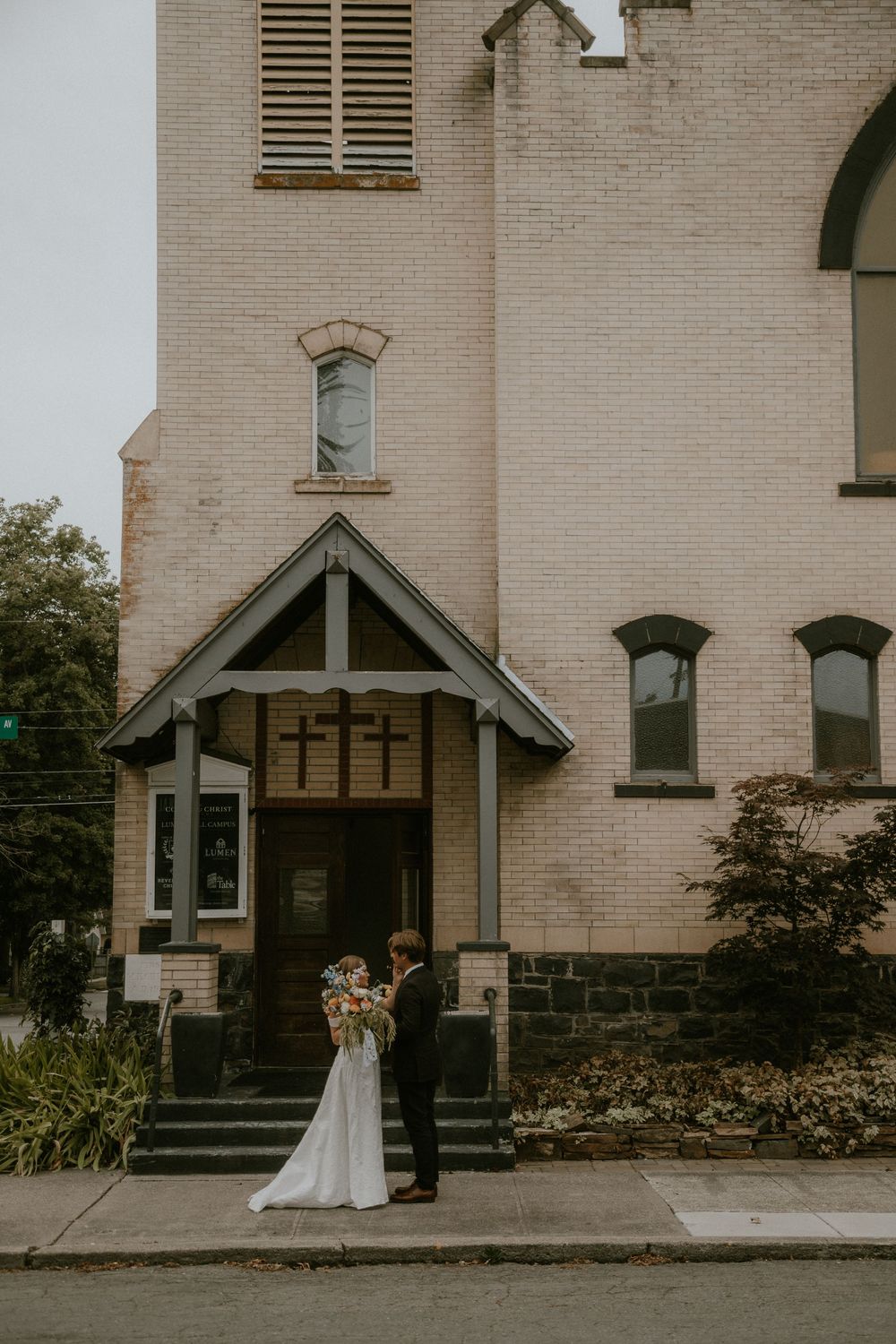 Bride in a white gown and groom in a dark suit embrace in front of a historic brick church entrance with a cross on the door.