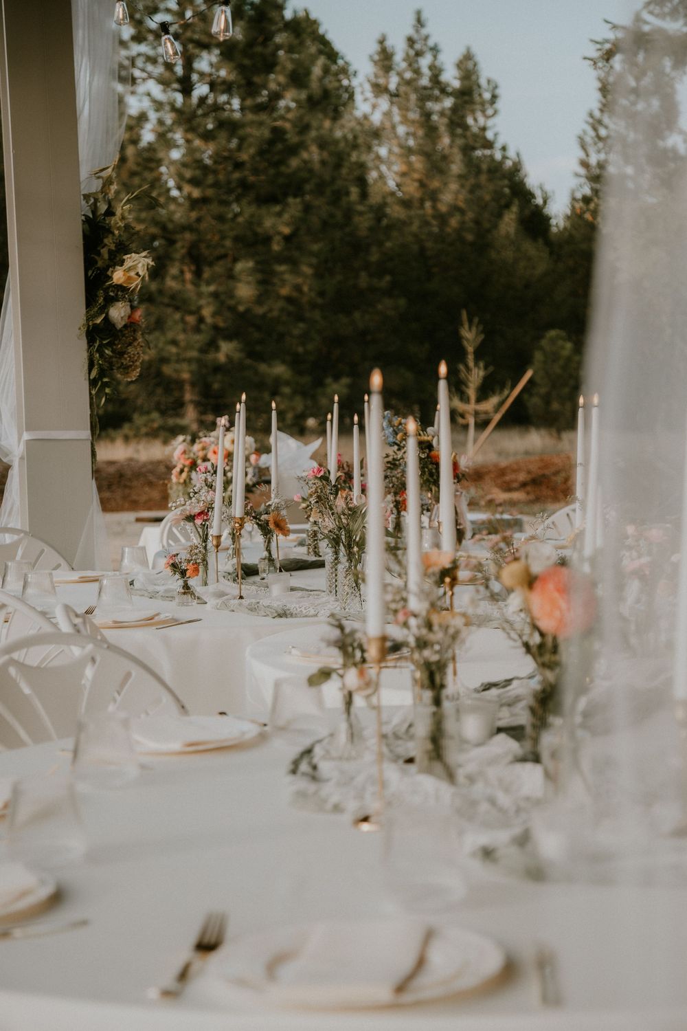 Outdoor wedding reception table with white linens, tall taper candles, peach floral arrangements, and place settings beside a pine forest.
