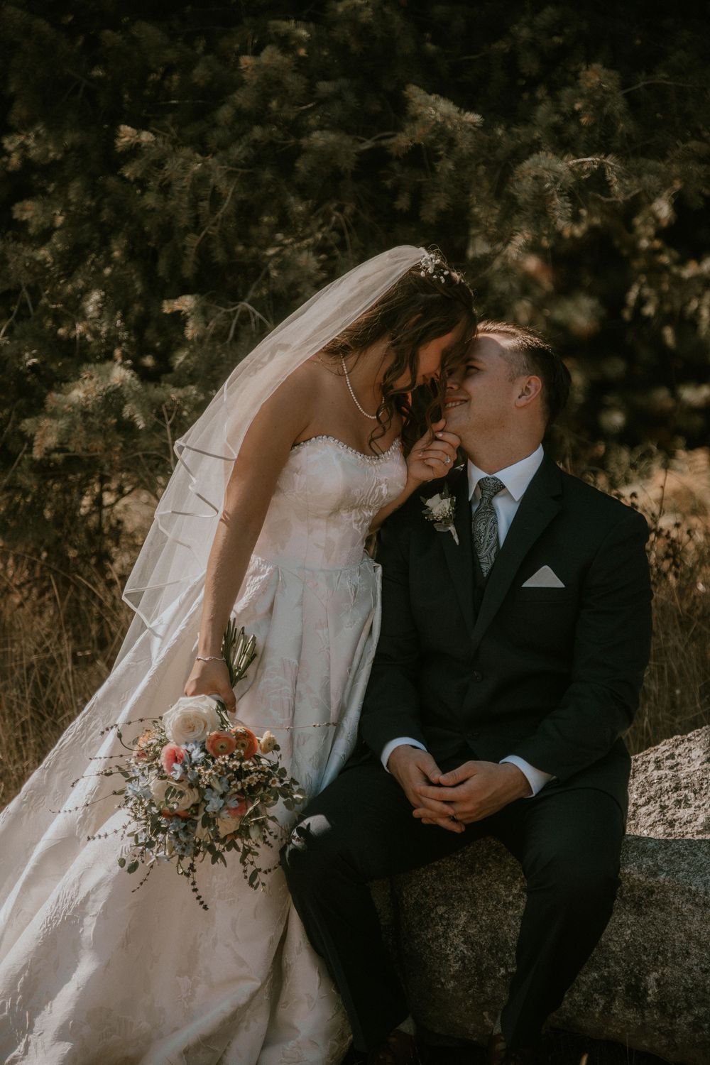 Bride in a white gown and veil leans in to kiss a seated groom in a dark suit, holding a floral bouquet outdoors.