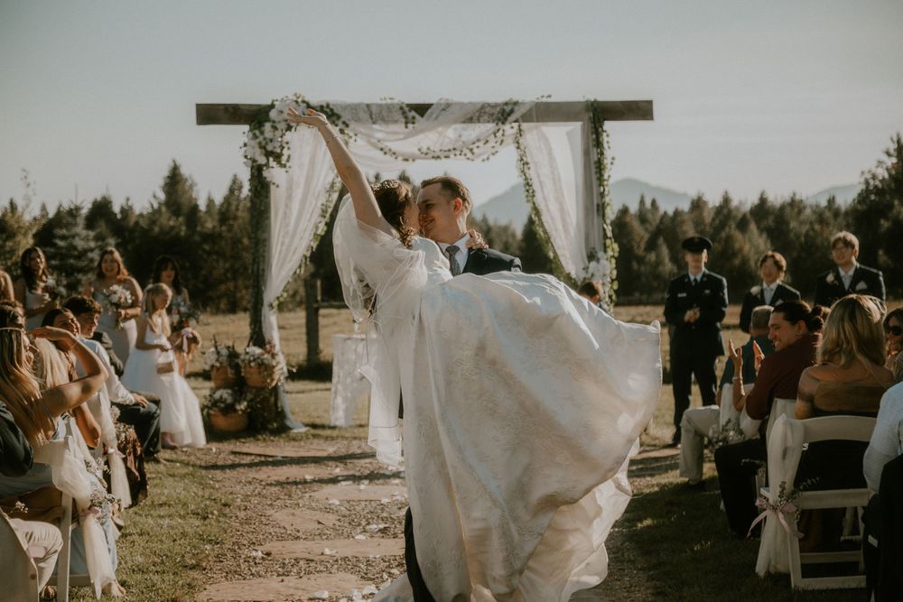 Bride in a flowing white gown kisses her groom under a draped wooden arch during an outdoor wedding ceremony as guests watch.