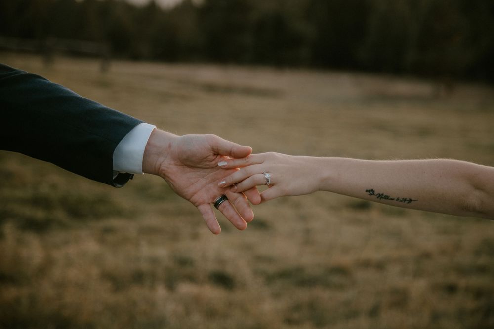 Close-up of a groom in a dark suit holding a bride's hand with wedding rings and a small arm tattoo in a grassy field.