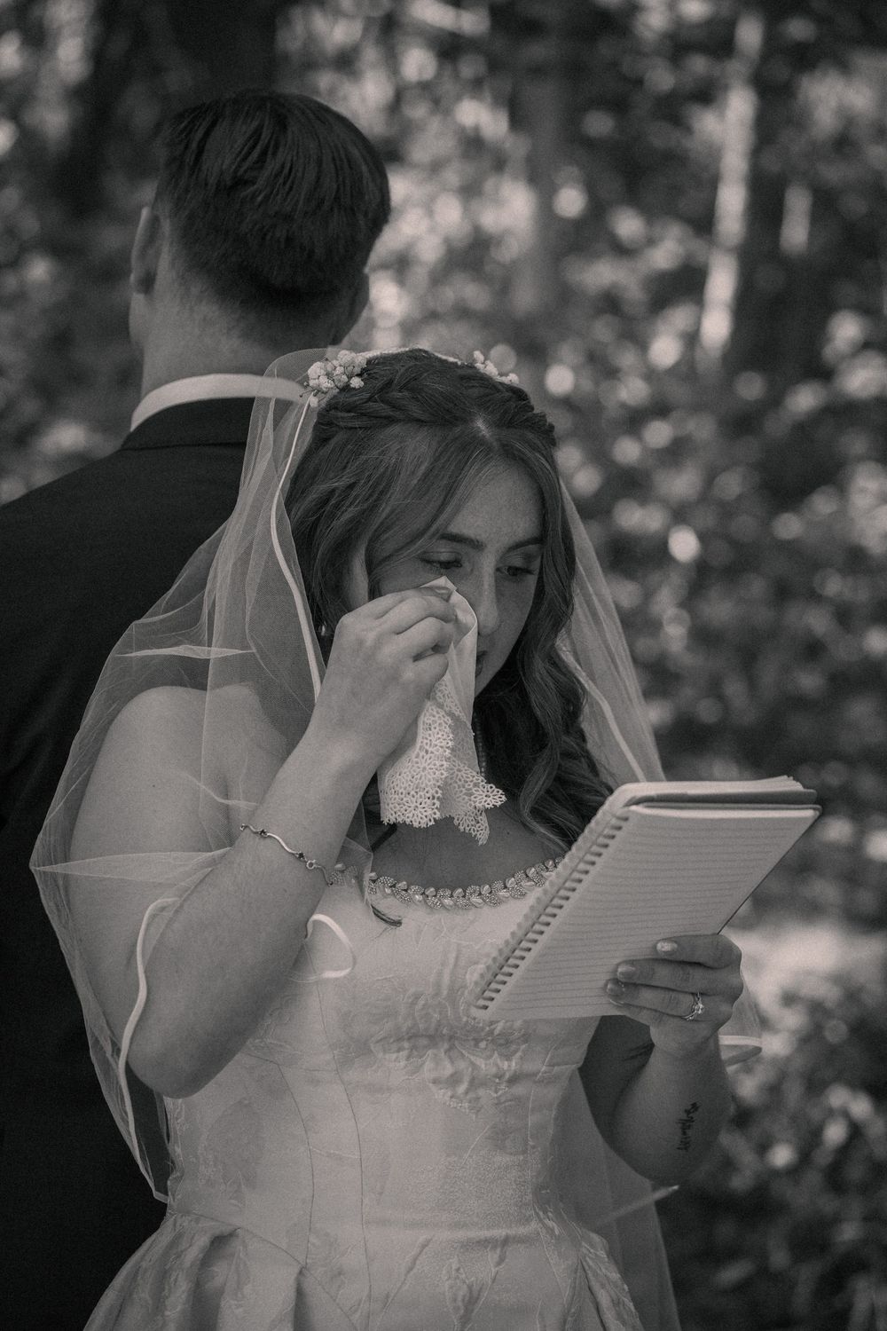 Black and white photo of a tearful bride in a veil dabbing her eyes with a handkerchief while reading vows from a notebook outdoors.
