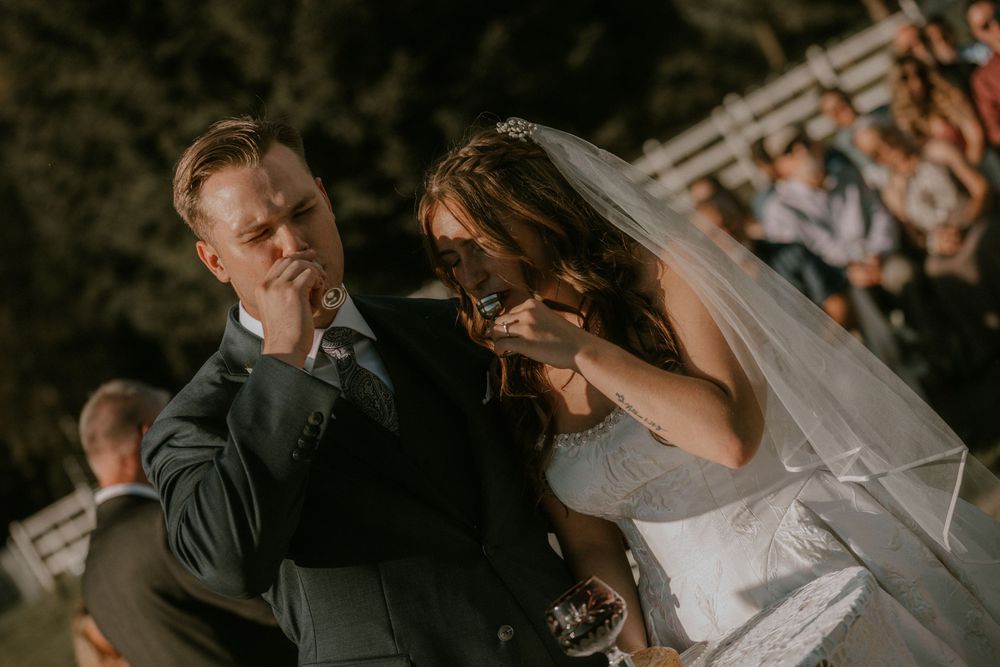 Bride in a white veil and groom in a gray suit share an emotional moment during an outdoor wedding ceremony as guests look on.