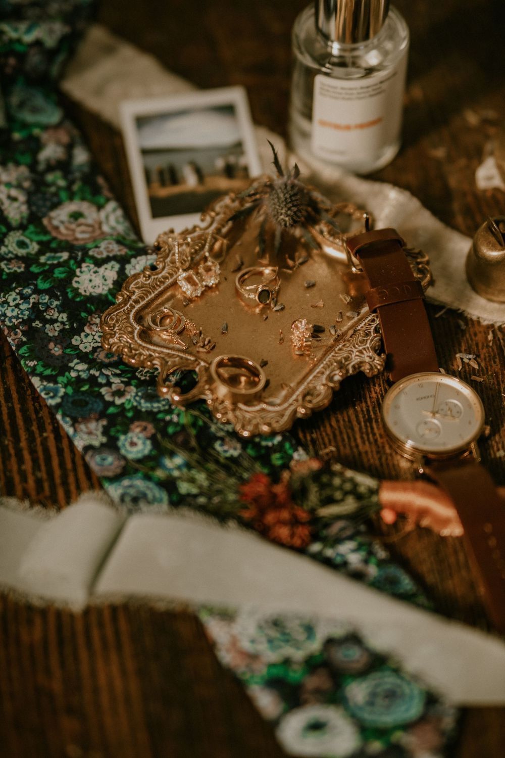 Gold jewelry tray with rings, a leather watch, polaroid photo, and perfume bottle on a floral fabric and wooden surface.