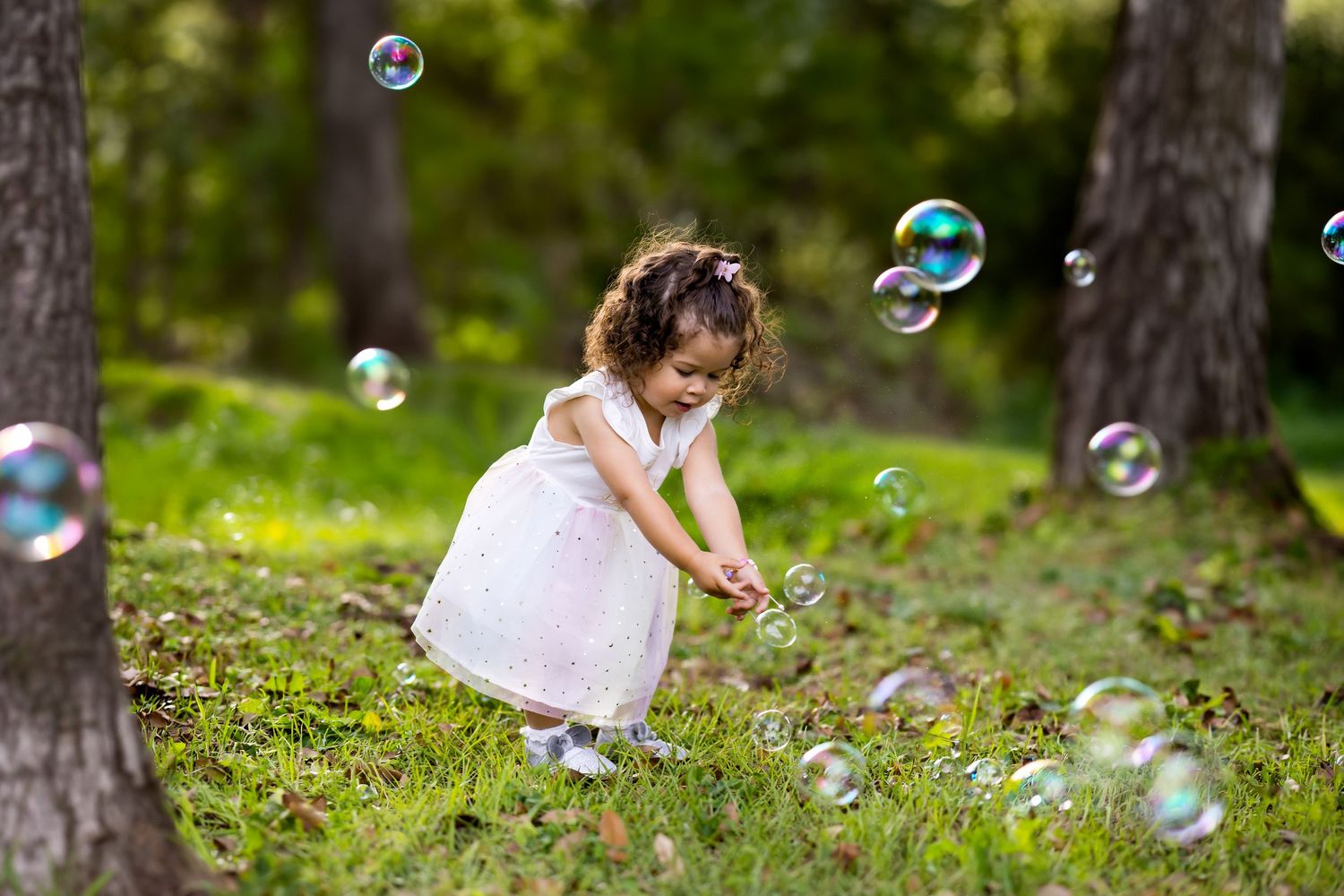 A young girl in a white dress plays with soap bubbles in a lush green park surrounded by trees.