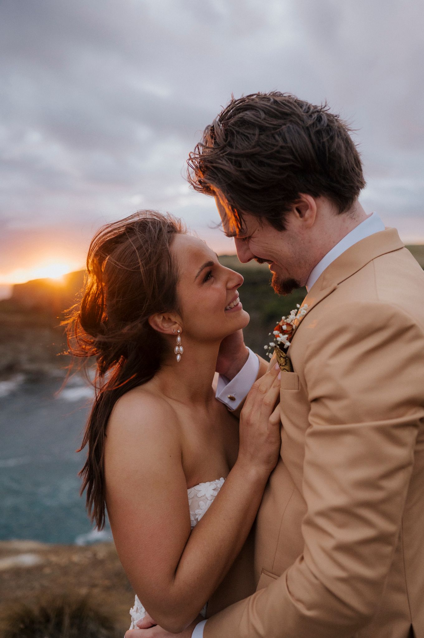 Bride and groom share a romantic sunset moment on coastal cliffs, smiling at each other in wedding attire.