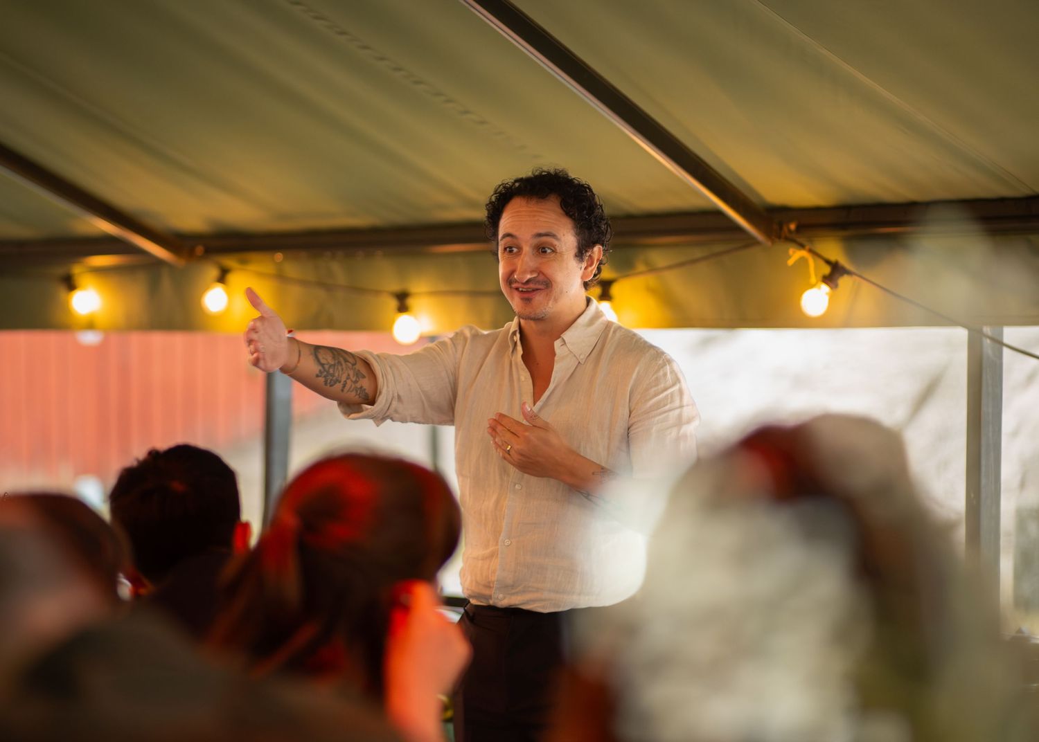 A tattooed man in a white shirt gestures enthusiastically while speaking under a tent at an event.