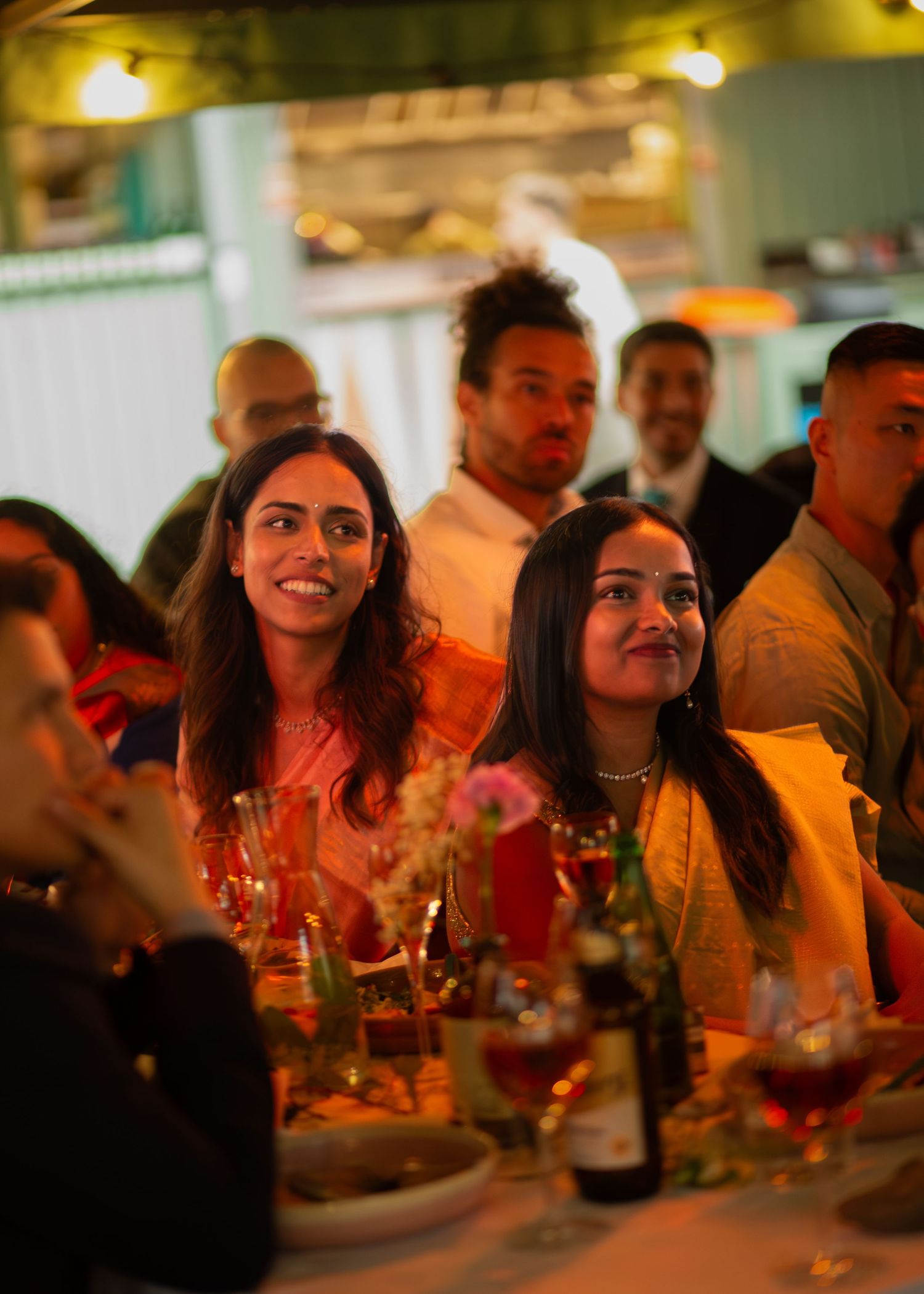 Guests smiling and enjoying a festive dinner event, seated at a decorated table with drinks and flowers. Bröllopsmiddag på Kajsa i Parken
In