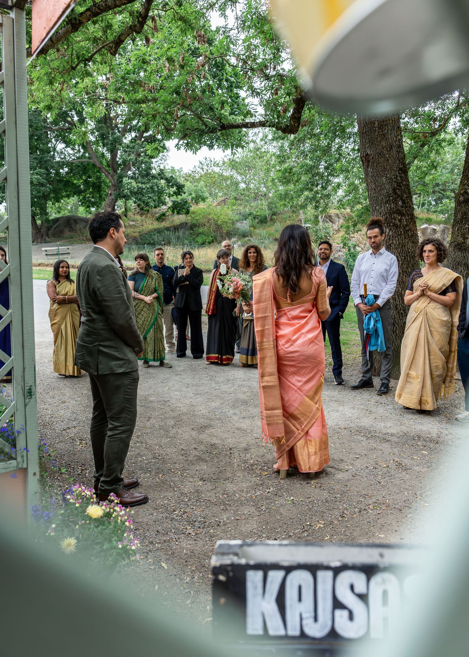 Indian wedding guests in traditional sarees gather outdoors near trees as a couple faces each other in a ceremonial moment.
