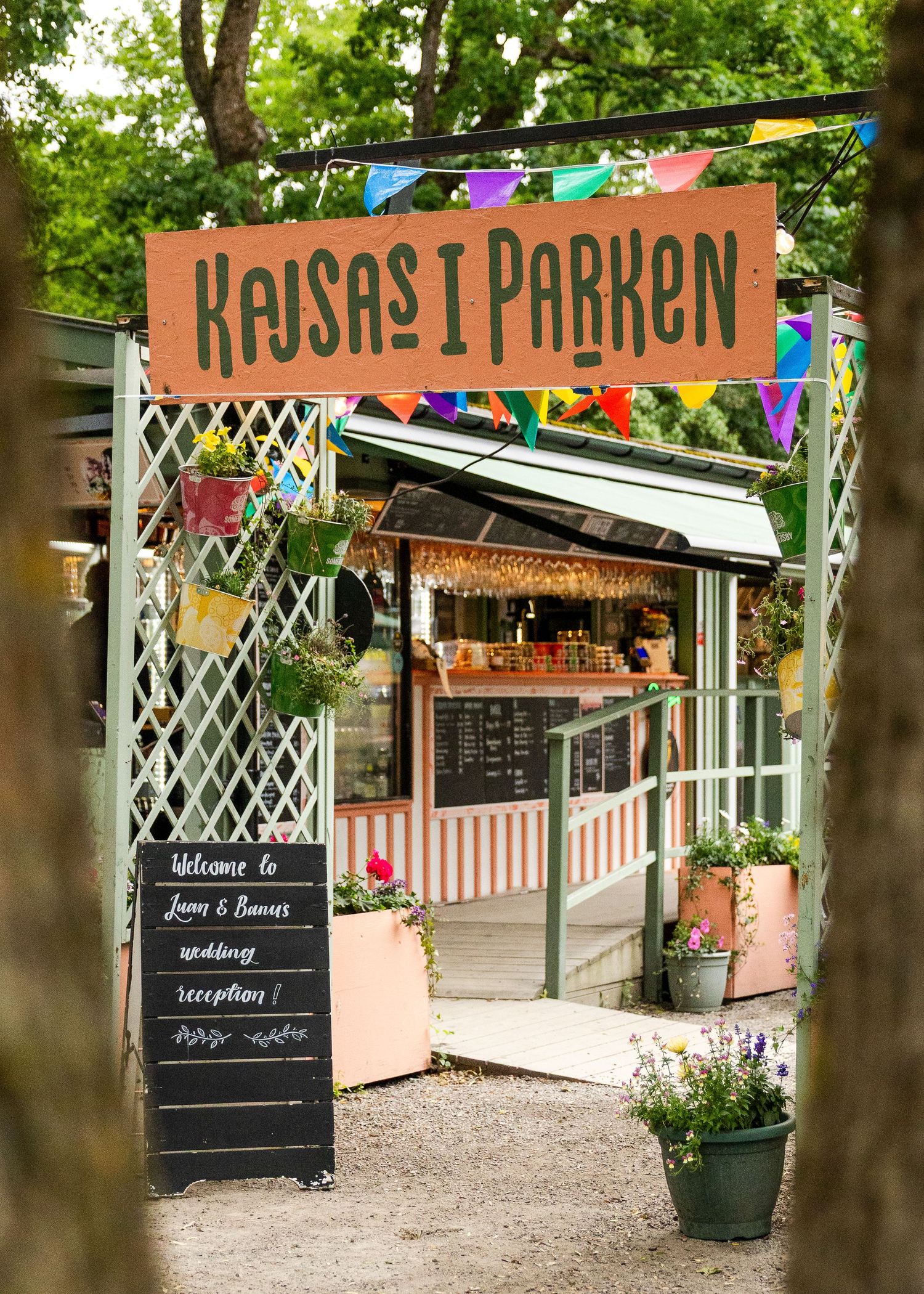 Kajsas i Parken outdoor cafe entrance with colorful bunting, welcome chalkboard sign, and flower decorations in a lush garden setting.