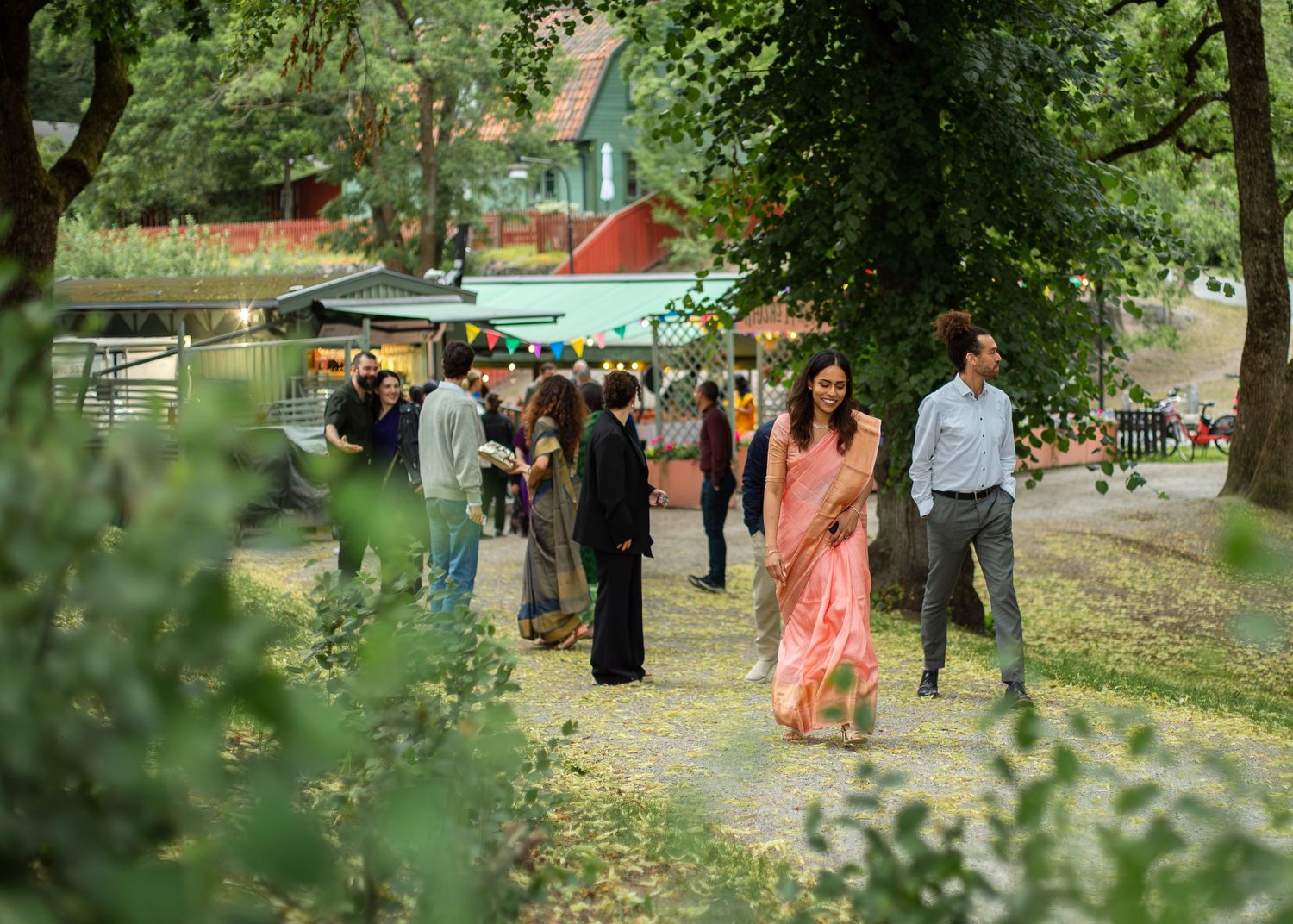 People in elegant attire, including a woman in a pink saree, stroll through a lush green outdoor garden event.