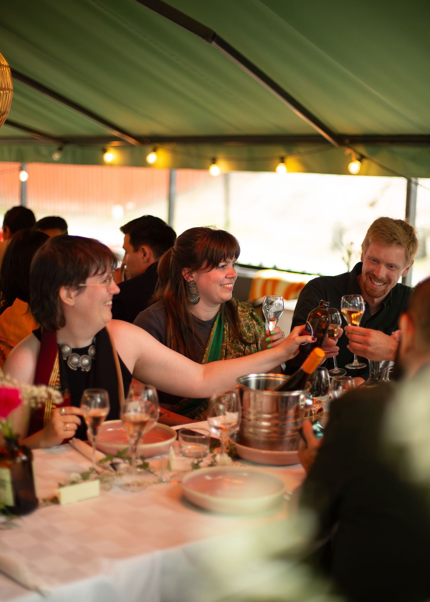 Guests laughing and toasting with wine glasses at an elegant outdoor dinner party under a green tent with string lights. Kajsa i Parken