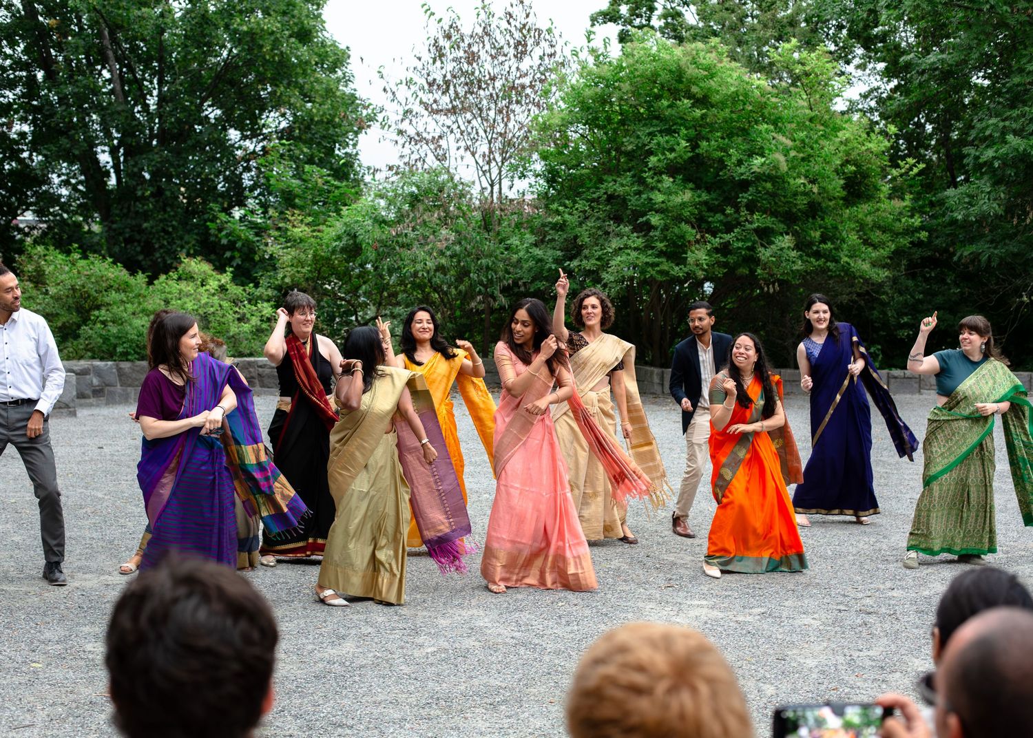 Women in colorful traditional Indian sarees perform a dance outdoors at a cultural celebration surrounded by lush green trees.