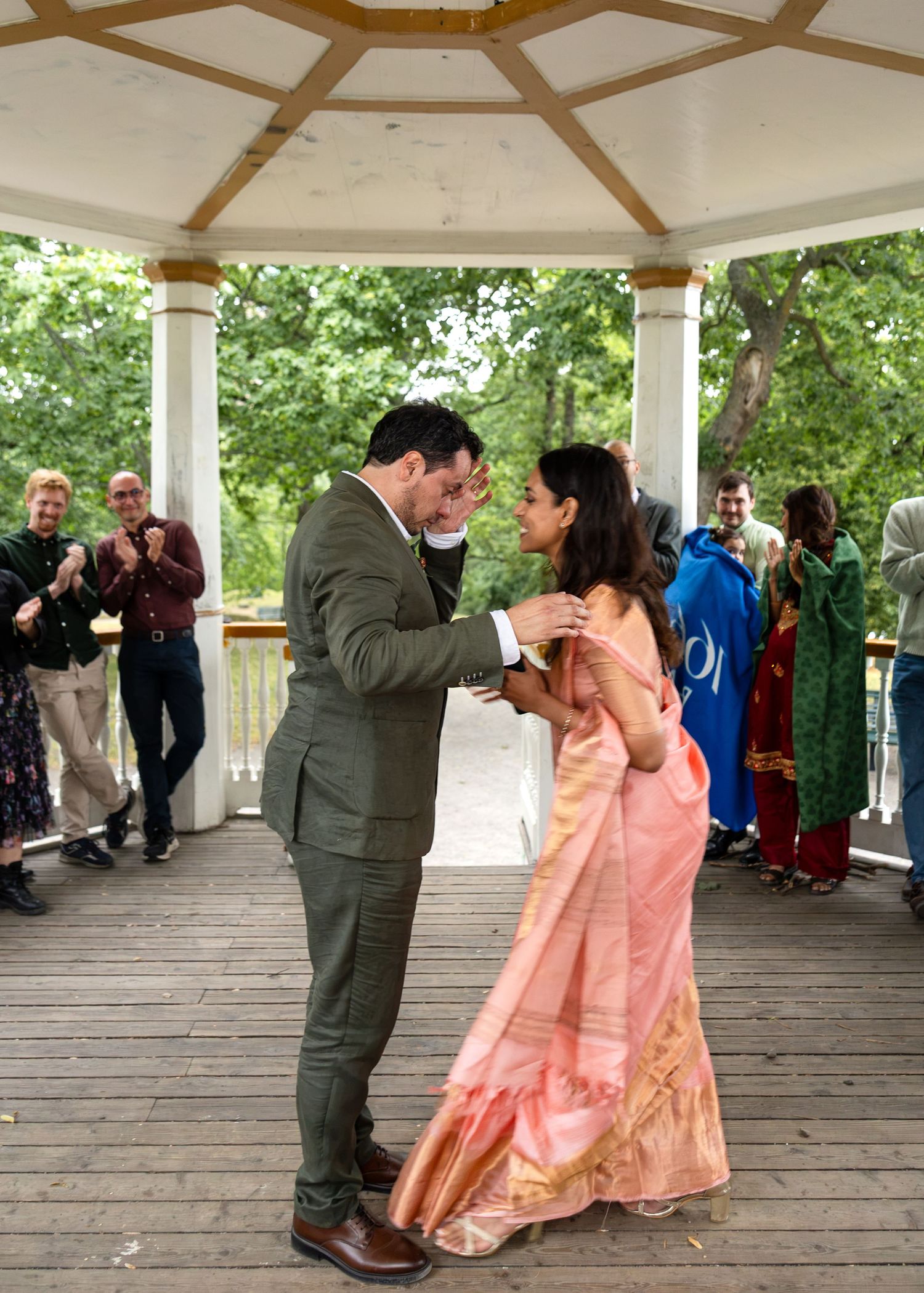 Couple sharing first dance in a gazebo, bride in pink saree, groom in green suit, guests applauding around them.