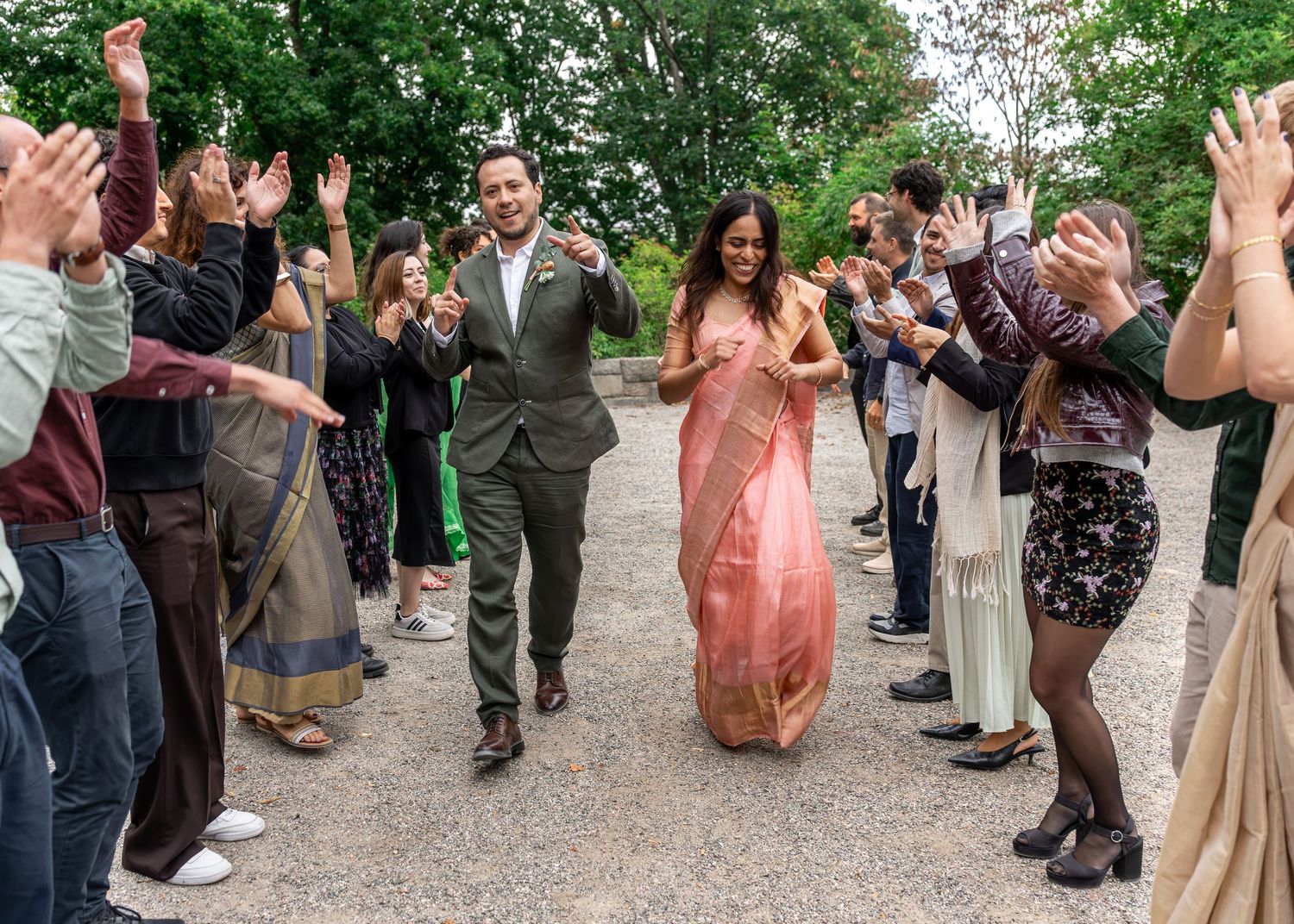 Couple in wedding attire walks through cheering guests outdoors, bride in pink saree, groom in gray suit. Bröllopsmiddag på Kajsa i Parken