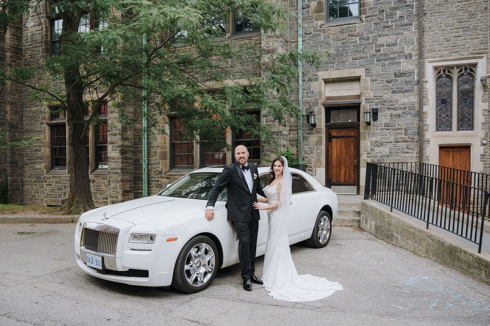 Bride and groom posing beside a white Rolls-Royce in front of a historic stone church on their wedding day.
