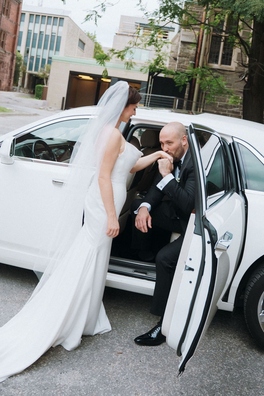 Groom in tuxedo kisses bride's hand while seated in white wedding limousine, bride in white gown and veil stands beside car.