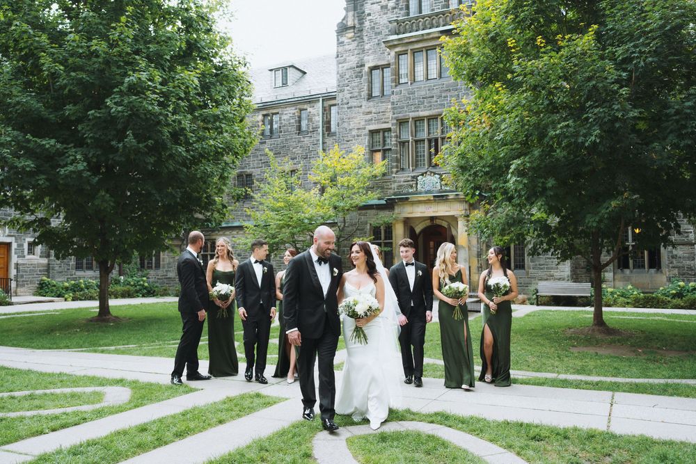 Bride and groom walk together with wedding party in front of a stone mansion surrounded by lush green trees.