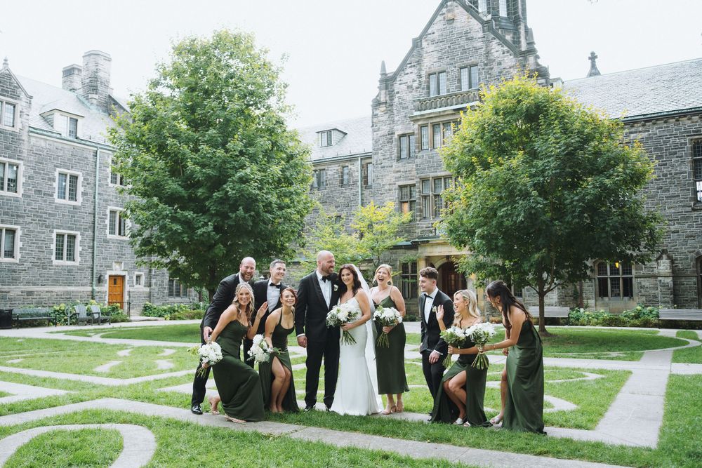 Wedding party poses in elegant stone courtyard, bridesmaids in green gowns flanking bride and groom.