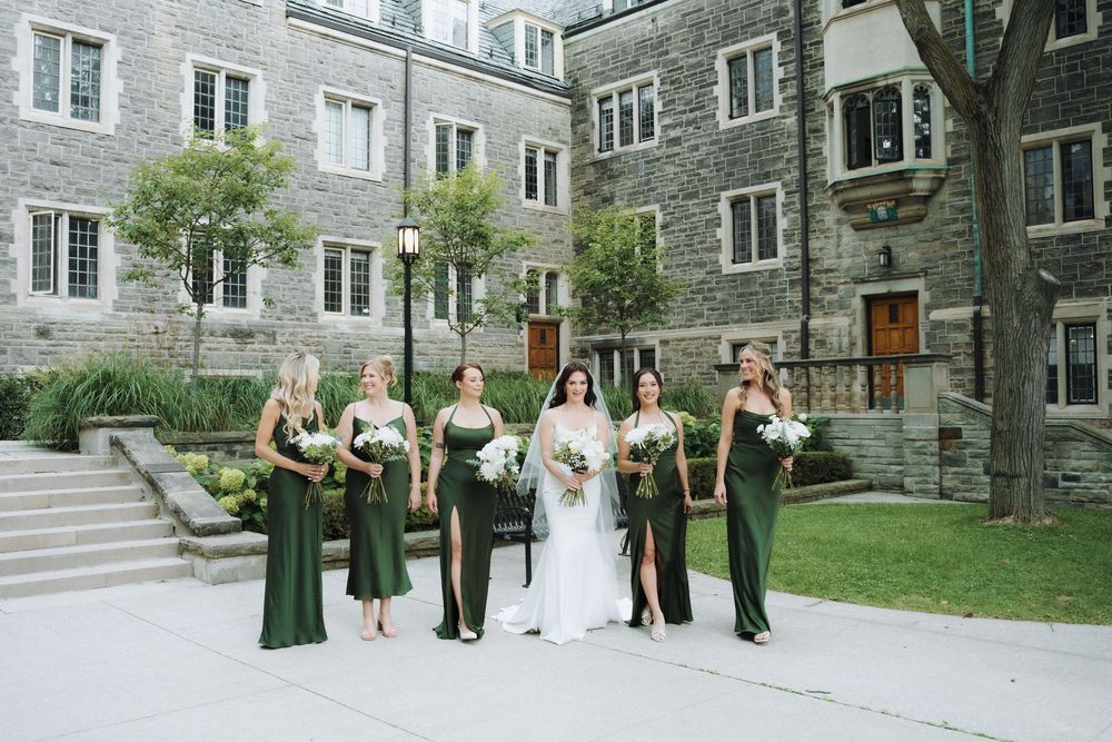 Bride in white gown with five bridesmaids in emerald green dresses holding white bouquets outside a stone building.
