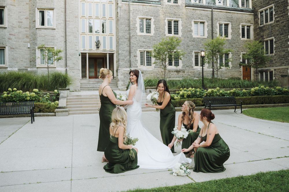 Bride in white gown with bridesmaids in green dresses outside a stone building on a sunny wedding day.