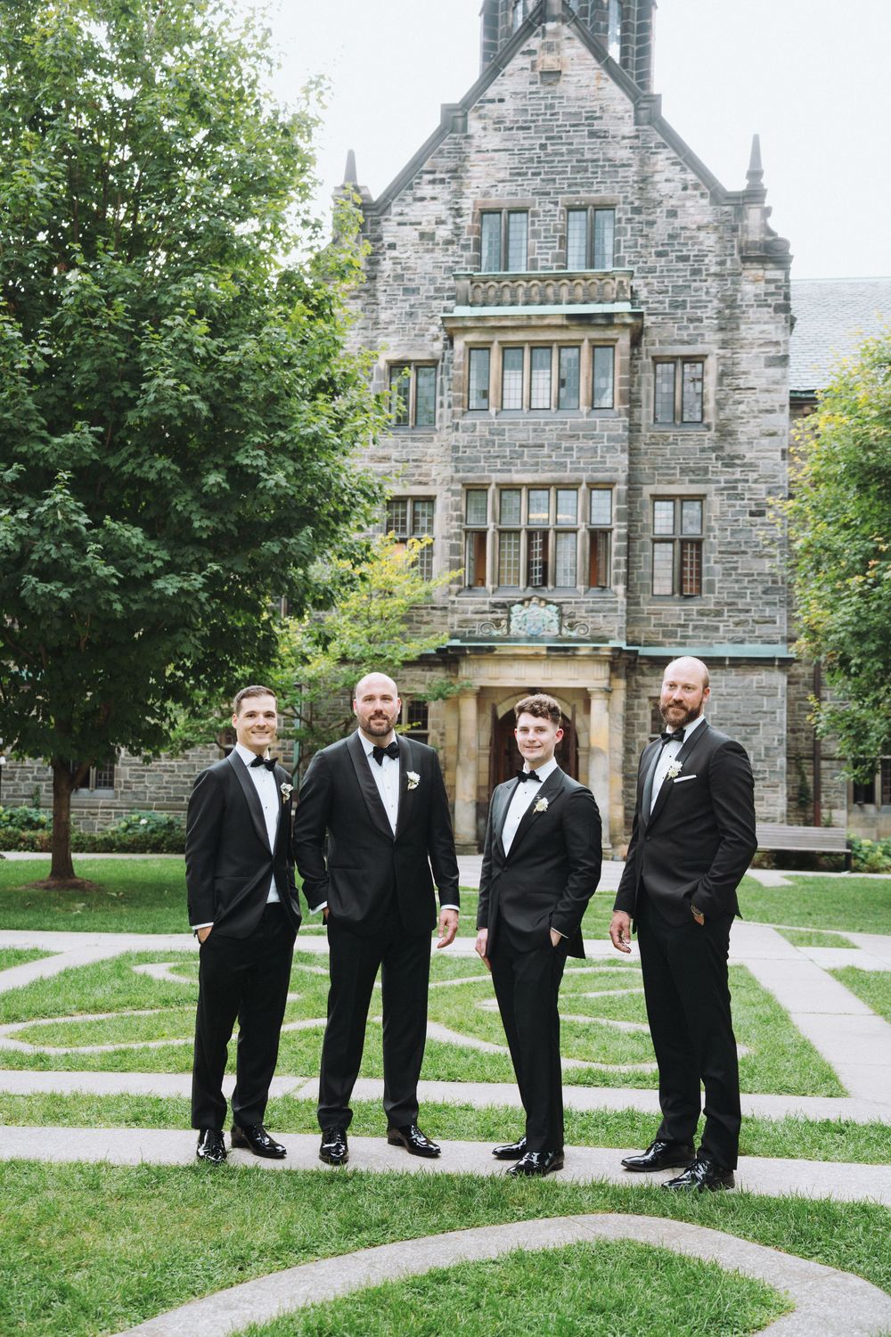 Four groomsmen in black tuxedos pose in front of a historic stone castle venue on a wedding day.