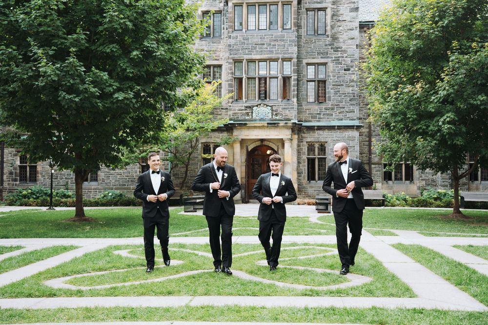 Four groomsmen in black tuxedos walk confidently across a manicured garden courtyard in front of a stone castle venue.
