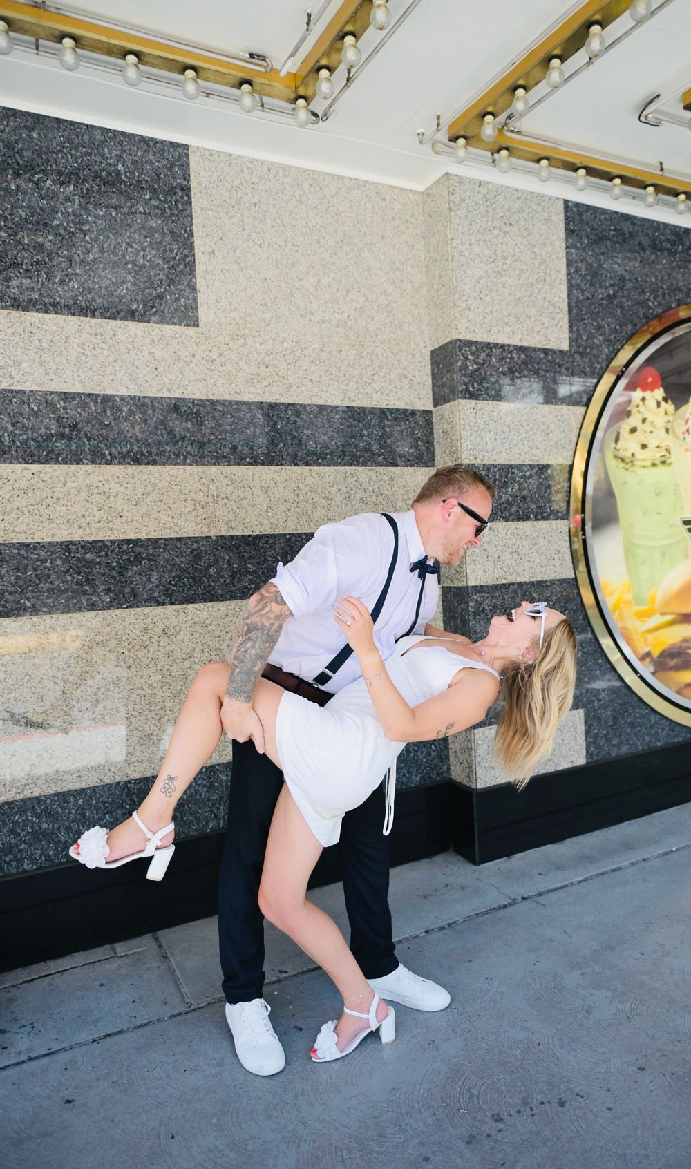 Groom dips bride in a romantic kiss outside a Las Vegas venue, both dressed in wedding attire with suspenders and white dress.