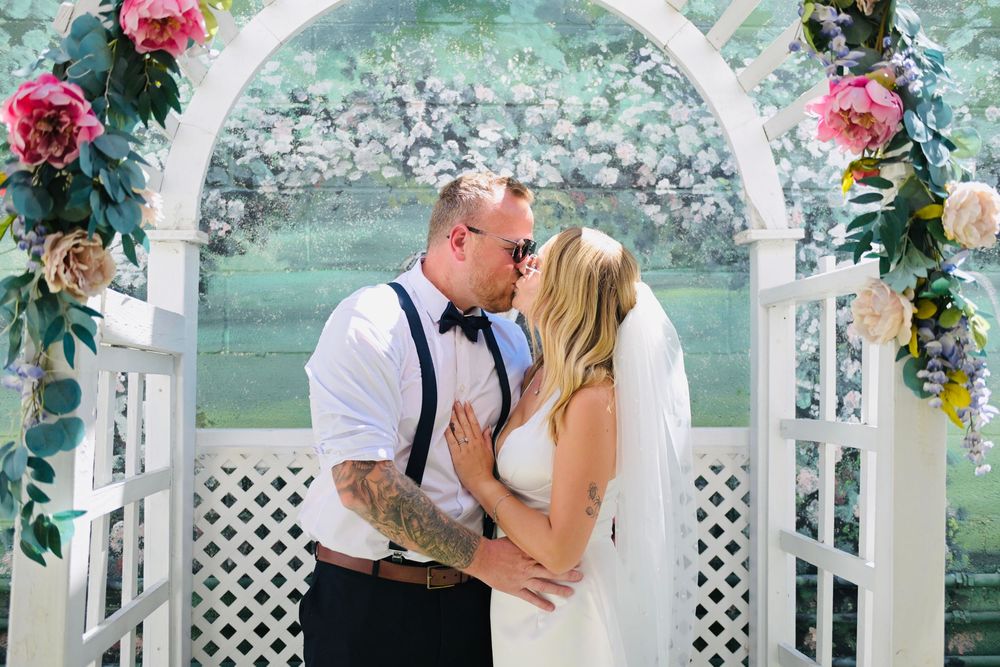 Bride and groom share a kiss under a floral-decorated white arbor at their wedding ceremony.