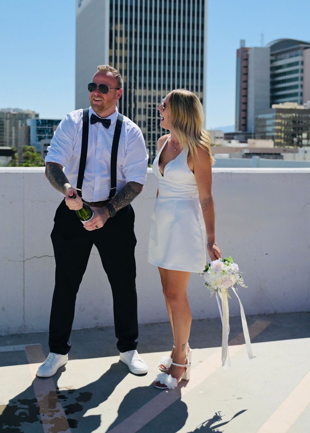 Couple in wedding attire on rooftop, groom in suspenders and bow tie, bride in white mini dress holding bouquet.
