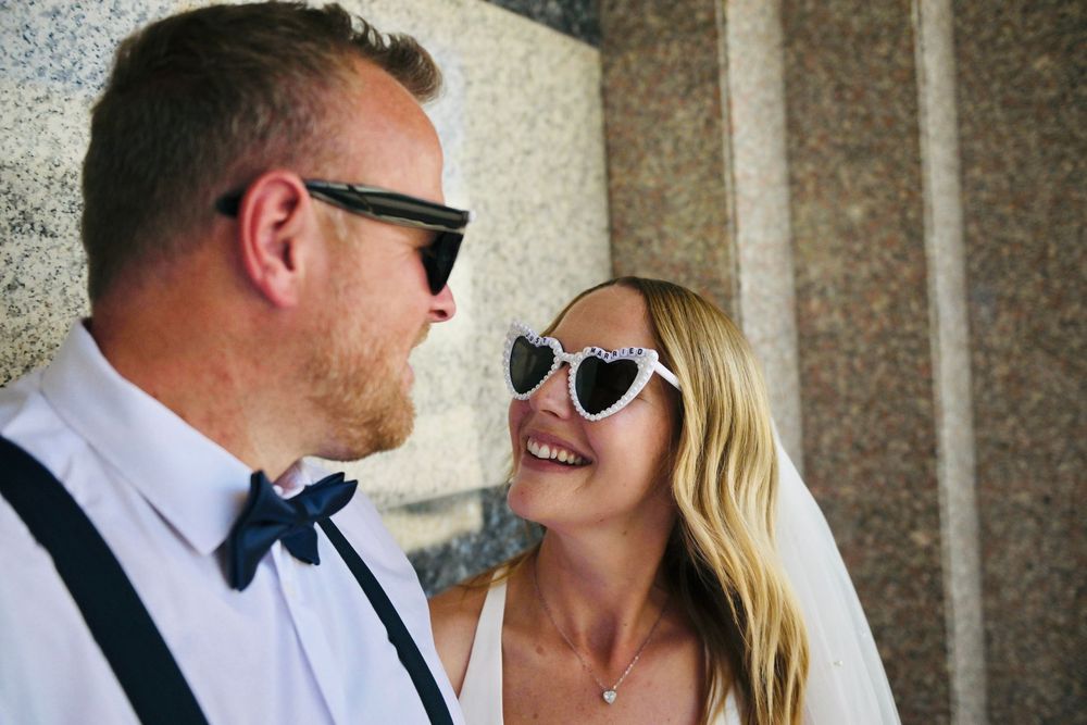 Bride in heart-shaped sunglasses and groom in bow tie and suspenders smiling at each other on their wedding day.