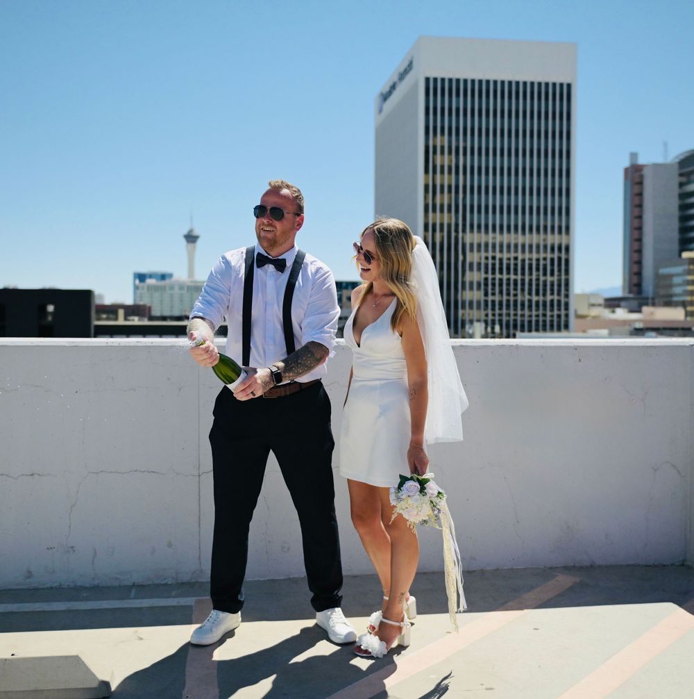 Newlyweds celebrate on a rooftop in Las Vegas, groom in suspenders holding champagne, bride in short white dress and veil.
