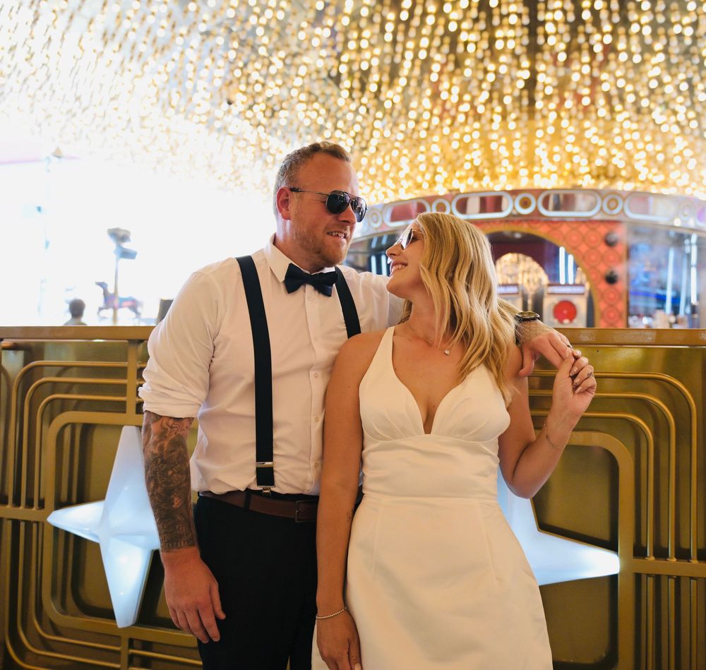 Couple in wedding attire smiling under sparkling chandelier in Las Vegas venue, groom in suspenders and bow tie.