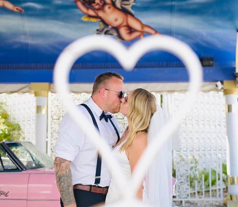 Newlyweds kissing framed by heart-shaped decoration at Las Vegas chapel with pink car in background.