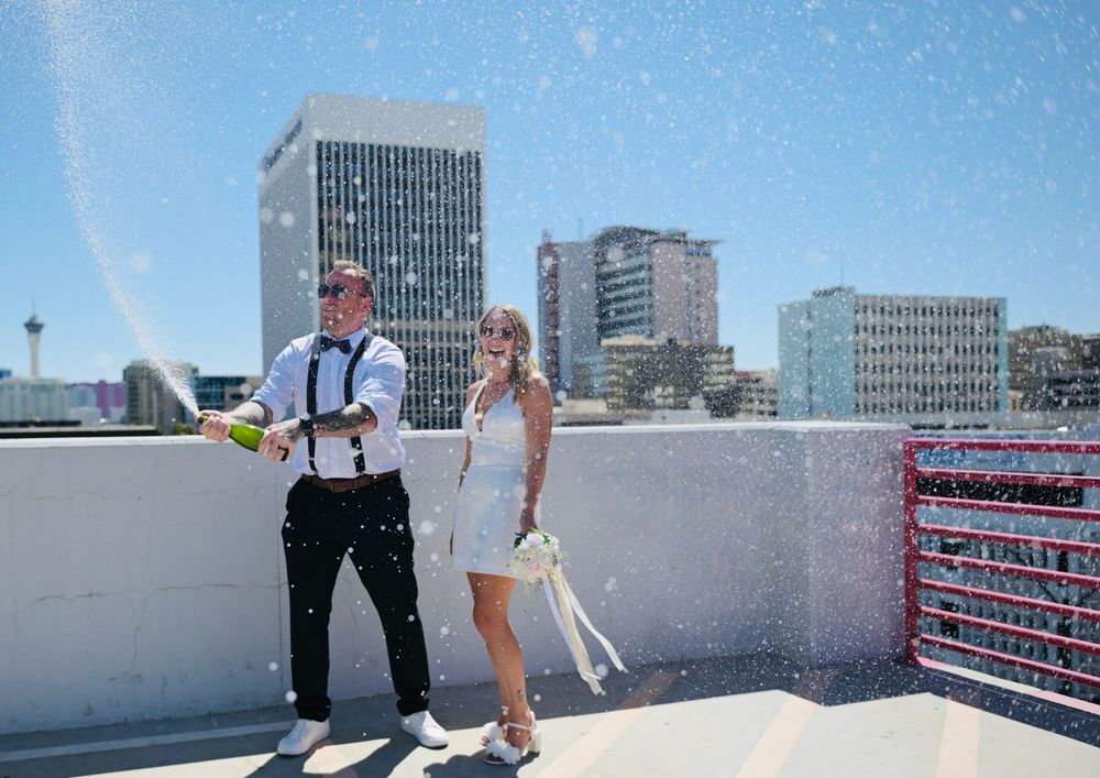 Newlyweds celebrating with champagne spray on a rooftop with Las Vegas city skyline in the background.
