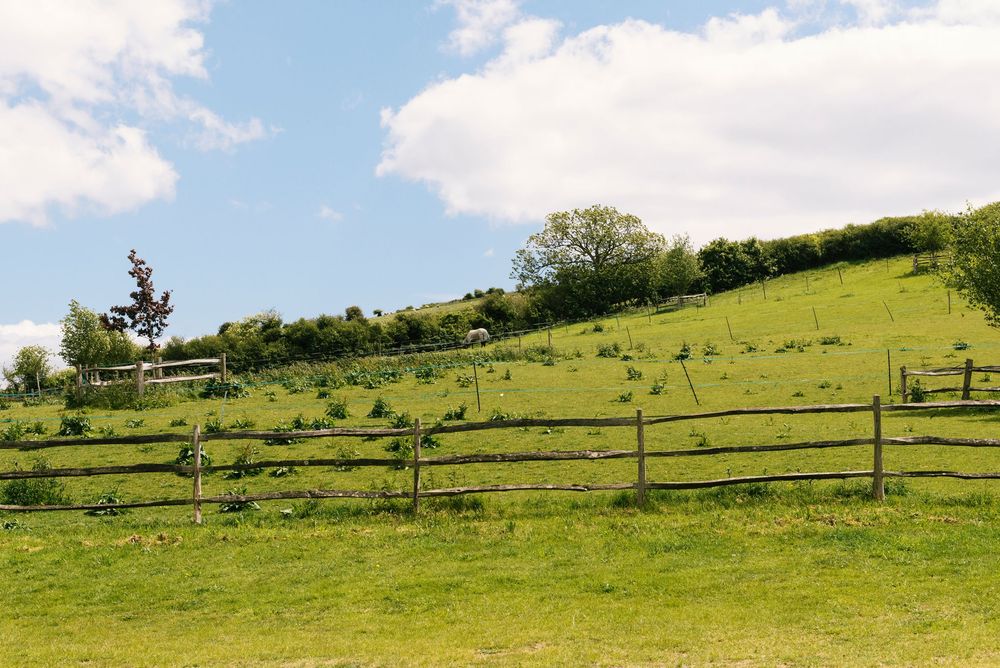 Lush green hillside farm with wooden post and rail fence under a partly cloudy blue sky.