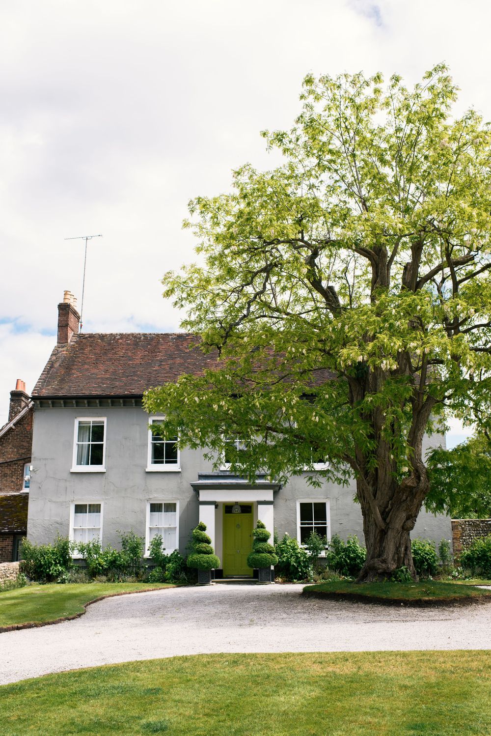 Charming grey English country house with large tree and green topiary by bright green front door.