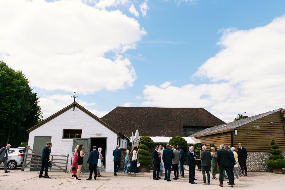 Wedding guests mingling outside a rustic barn venue under a blue sky with white clouds.