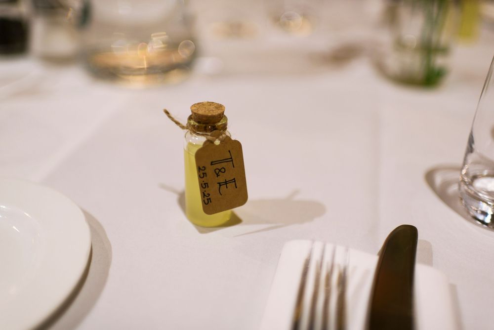 Small honey bottle wedding favor with cork stopper and kraft paper tag on white tablecloth at reception.