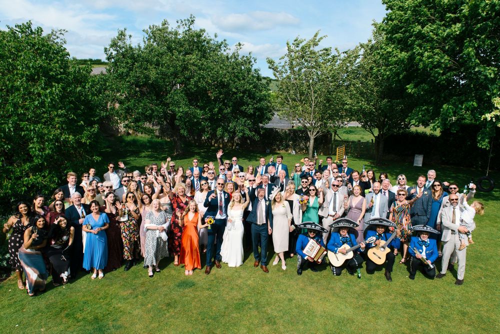 Large wedding group photo outdoors on green lawn with trees, bride and groom centered among smiling guests in colorful attire.