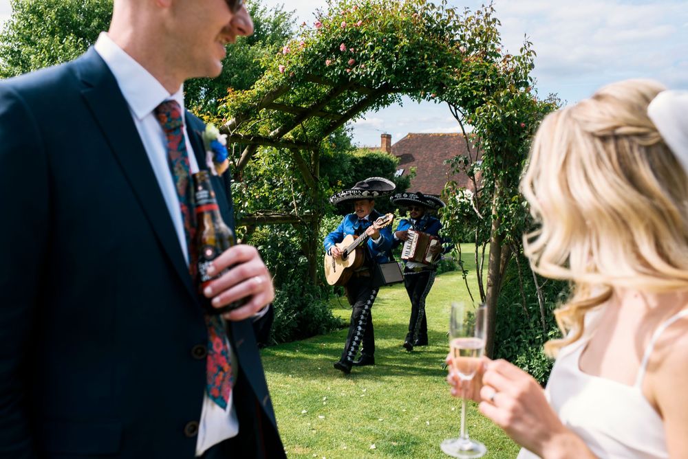 Mariachi band performs outdoors at a wedding reception while bride and groom enjoy champagne and beer in sunny garden.