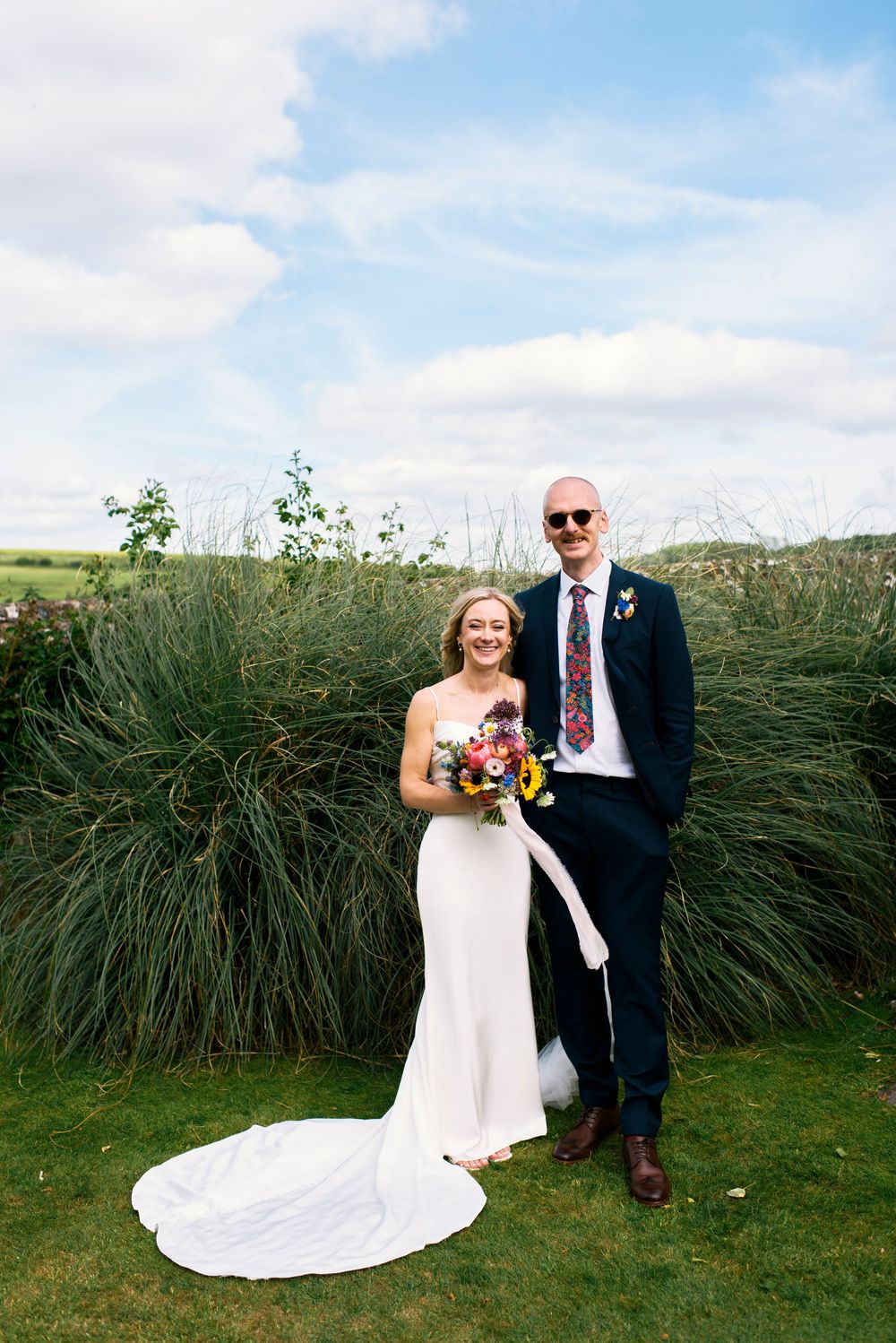 Bride and groom pose outdoors on green grass, she holds a colorful bouquet in a white gown, he wears a navy suit with floral tie.