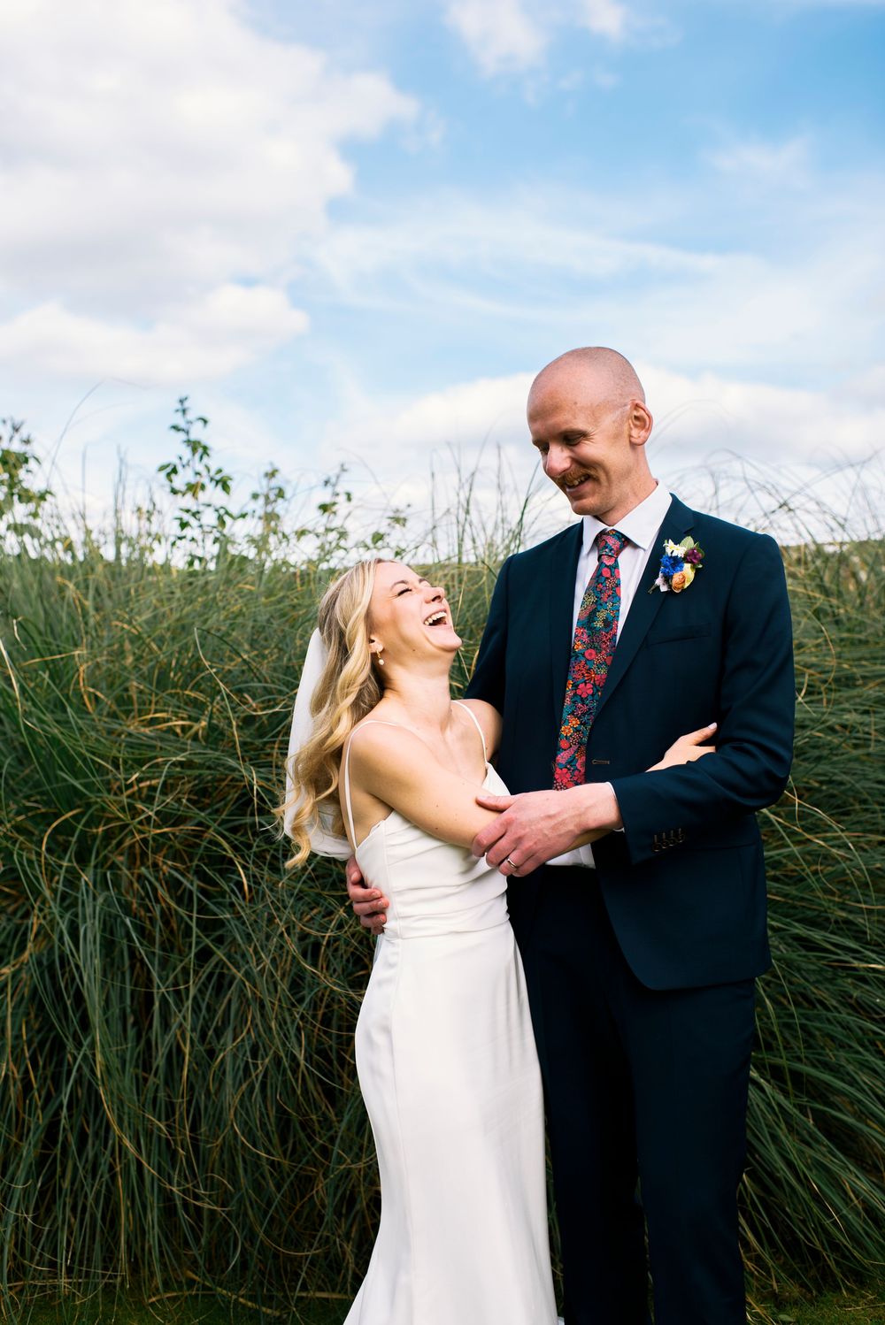 Laughing bride in white gown embraces groom in navy suit with floral tie outdoors against tall green grass.