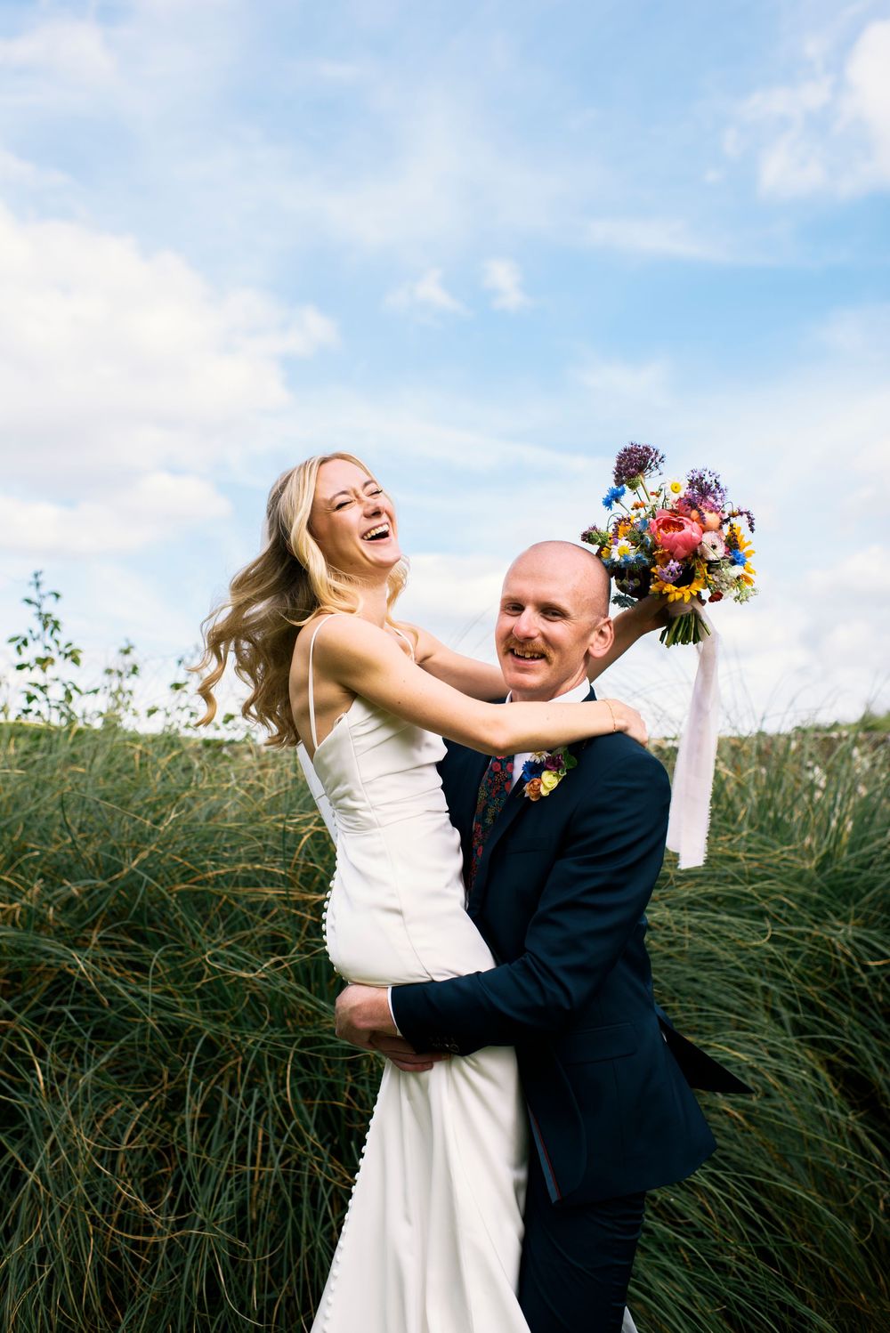 Joyful bride laughing as groom lifts her in a field, holding a colorful wedding bouquet under a blue sky.
