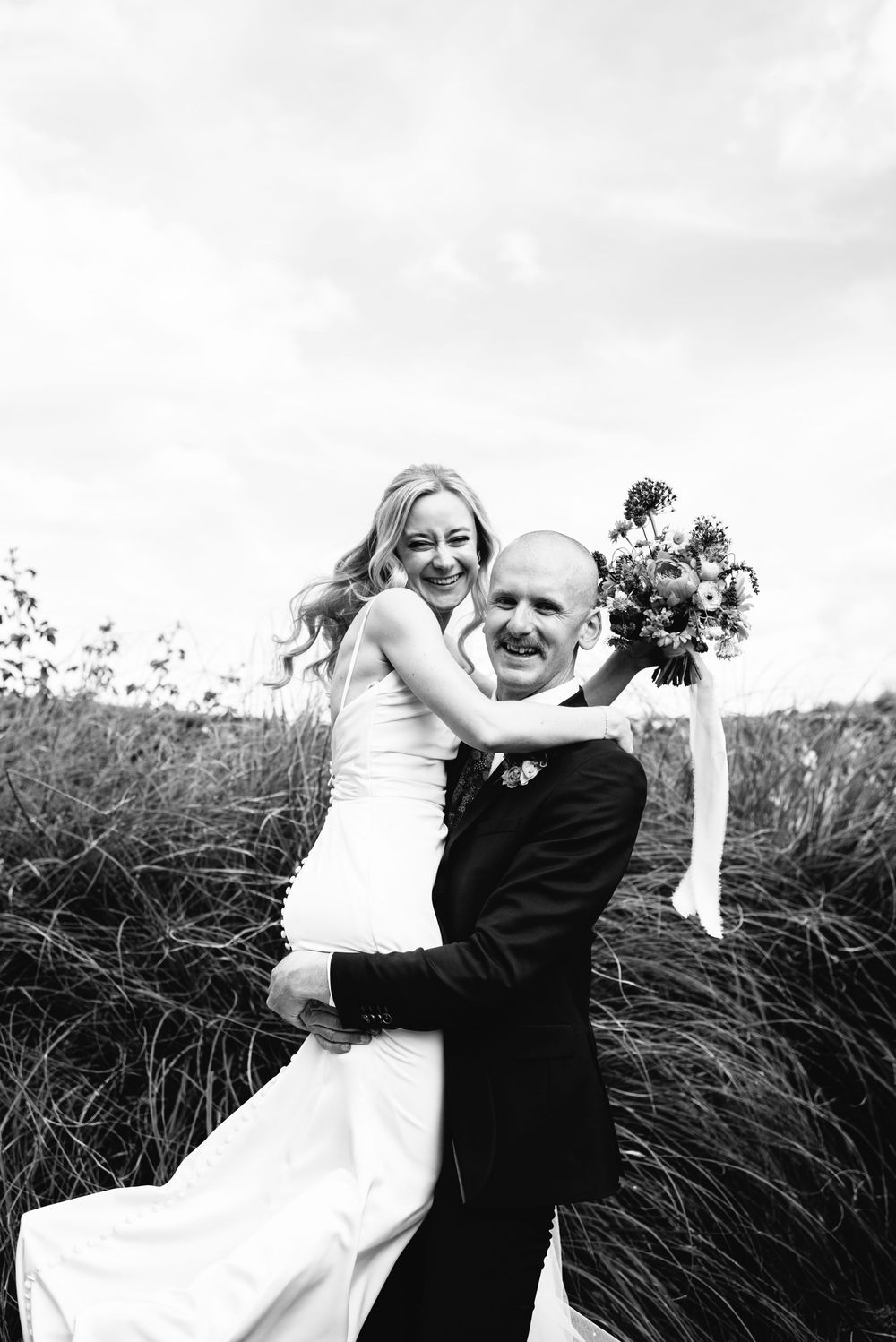 Black and white photo of a groom lifting his smiling bride in a field, both radiating joy on their wedding day.
