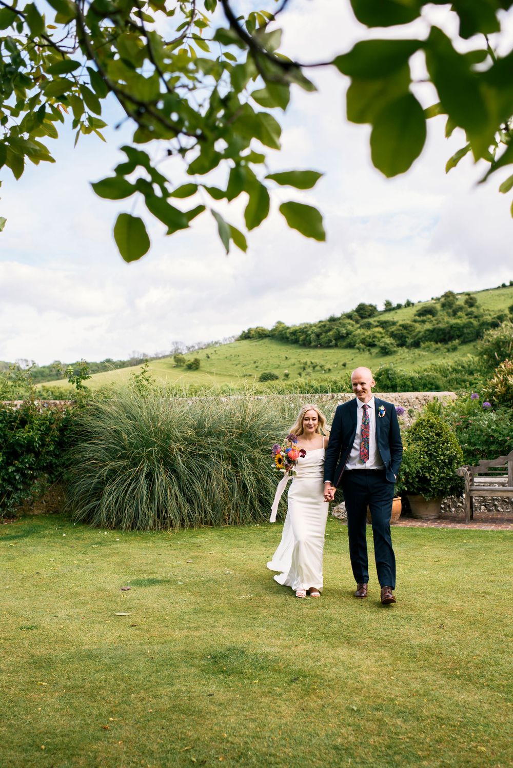 Bride and groom walking hand in hand across green lawn with rolling hills backdrop on their wedding day.