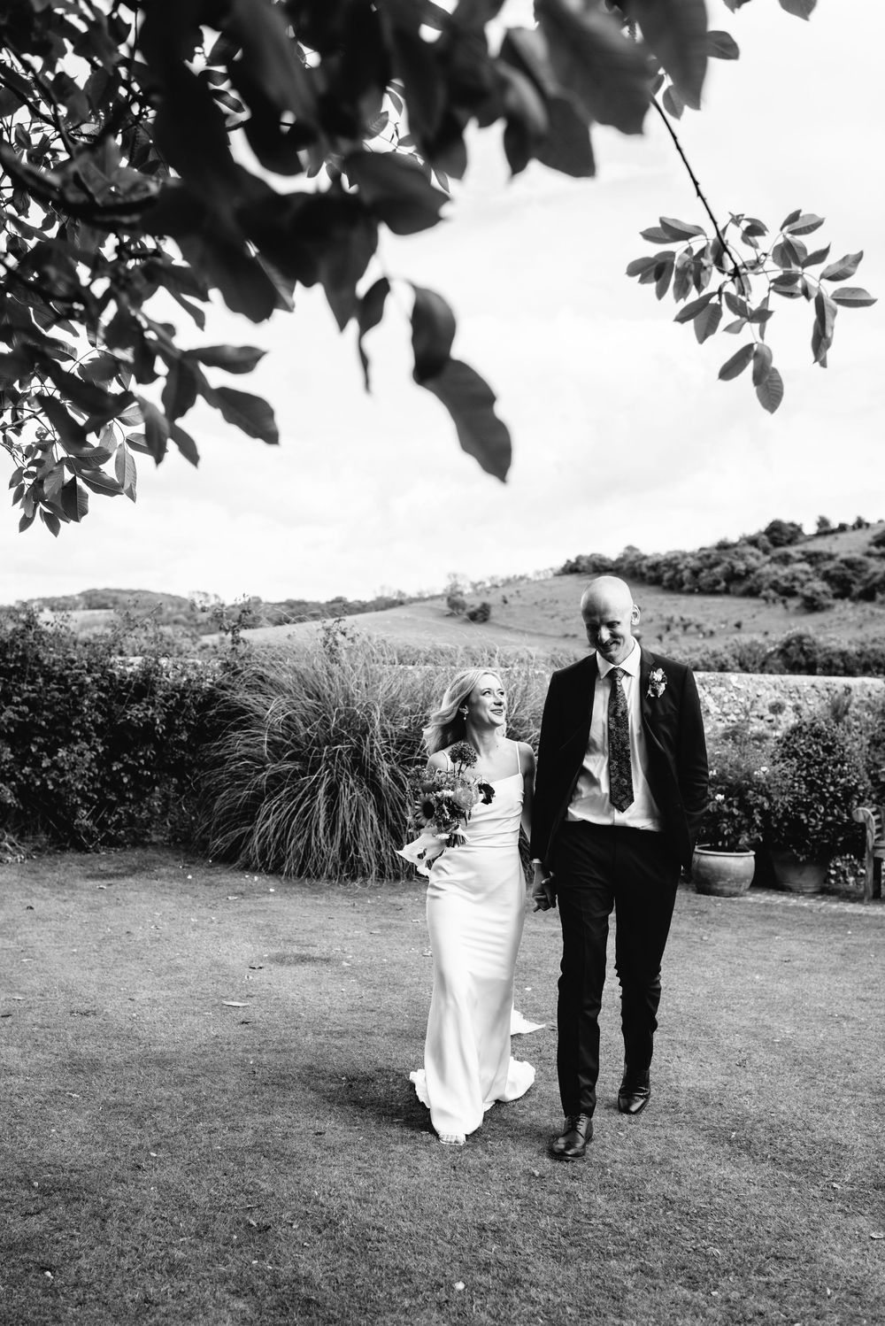 Black and white photo of bride and groom walking hand in hand across lawn with rolling hills backdrop.