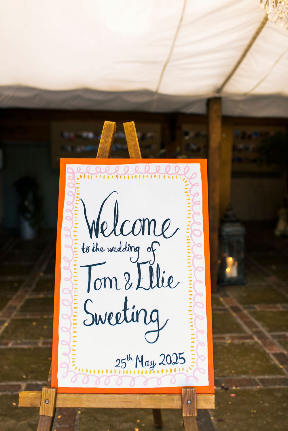 Wedding welcome sign on easel for Tom and Ellie Sweeting, 25th May 2025, with orange border and handwritten text.